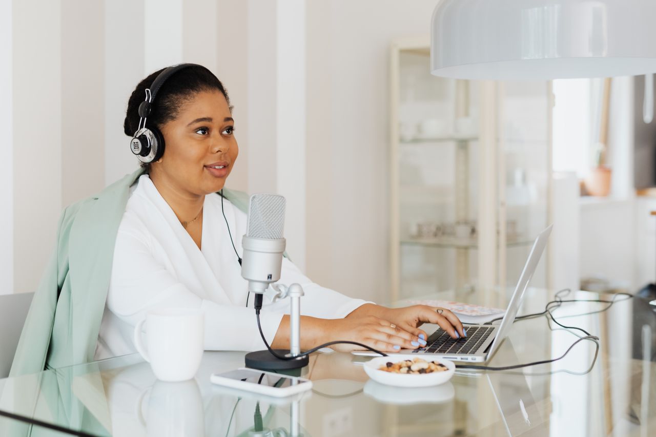 A woman working on a podcast in a modern home studio setup, using a laptop and microphone.